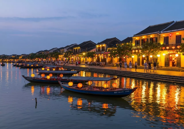 Khung cảnh phố cổ Hội An tuyệt đẹp lúc hoàng hôn, những chiếc đèn lồng rực rỡ chiếu sáng đường phố và sông Thu Bồn, thuyền gỗ truyền thống nhẹ nhàng trôi, du khách tản bộ dọc bờ sông, tạo nên không khí lãng mạn và sống động.